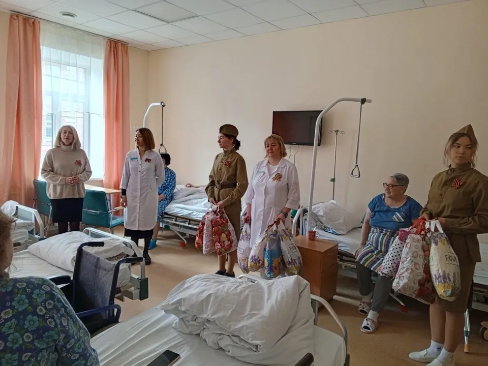 Женщины в форме сестёр милосердия поздравляют пожилого ветерана в палате госпиталя