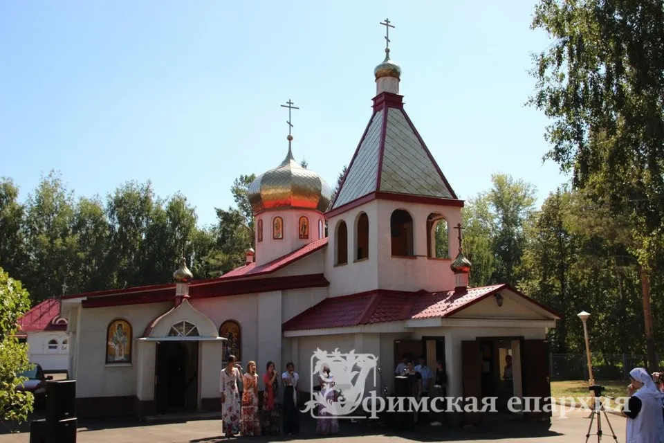 Небольшой храм с золочёными куполами, красной кровлей и звонницей летом, среди деревьев; у входа — небольшая группа прихожан