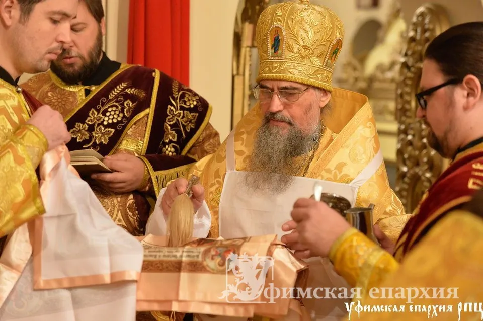Архиерей в золотом облачении благословляет каравай в интерьере храма, вокруг духовенство и прихожане