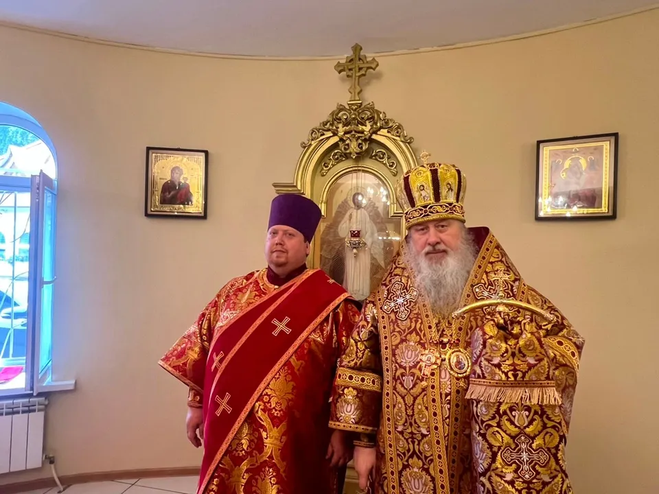 Диакон в красном облачении и фиолетовой камилавке рядом с митрополитом в полном облачении